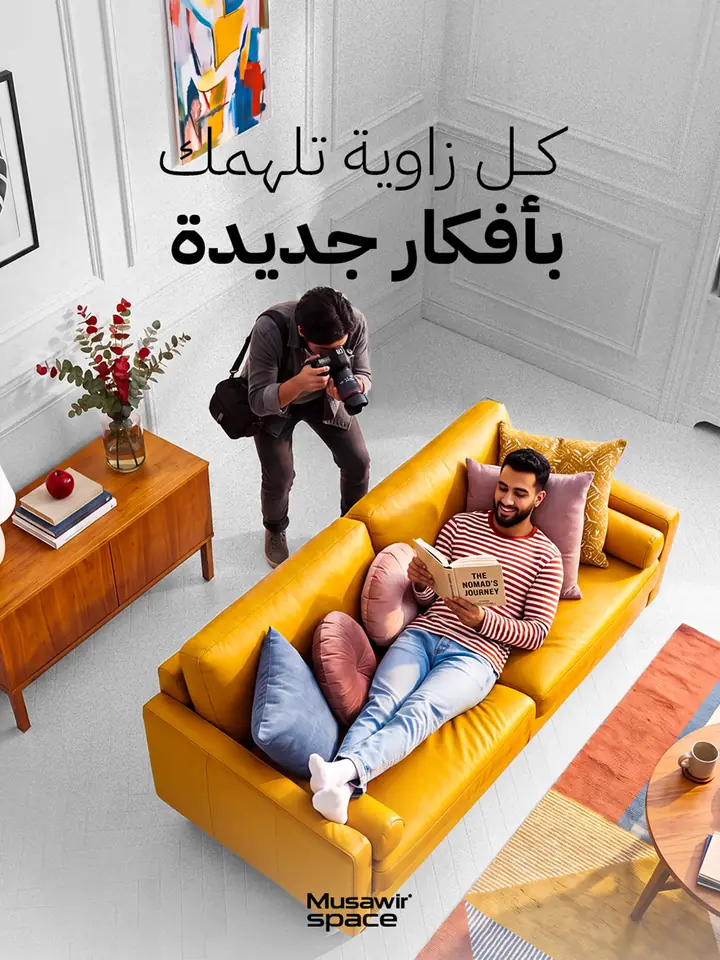 إعلان لمنصة حجز مواقع تصوير: مصور ينحني فوق أريكة صفراء يلتقط شابًا يقرأ كتابًا في شقة بيضاء